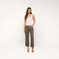A woman stands against a plain white background, wearing a white tank top, the Wanda Cropped Wide-Leg Jean in olive green with a tan belt, and tan heeled sandals for a touch of Parisian style.