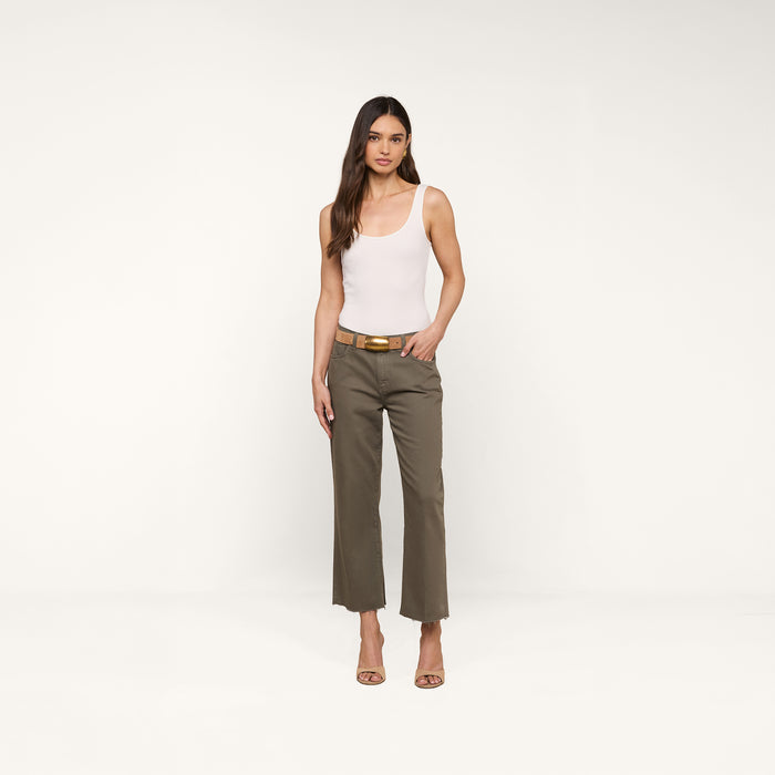 A woman stands against a plain white background, wearing a white tank top, the Wanda Cropped Wide-Leg Jean in olive green with a tan belt, and tan heeled sandals for a touch of Parisian style.