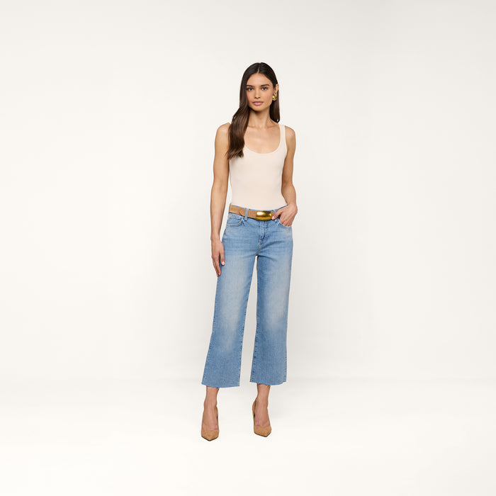 A woman stands against a plain background wearing a beige tank top, the Wanda Cropped Wide-Leg Jean in a light blue wash, a brown belt, and tan heels.