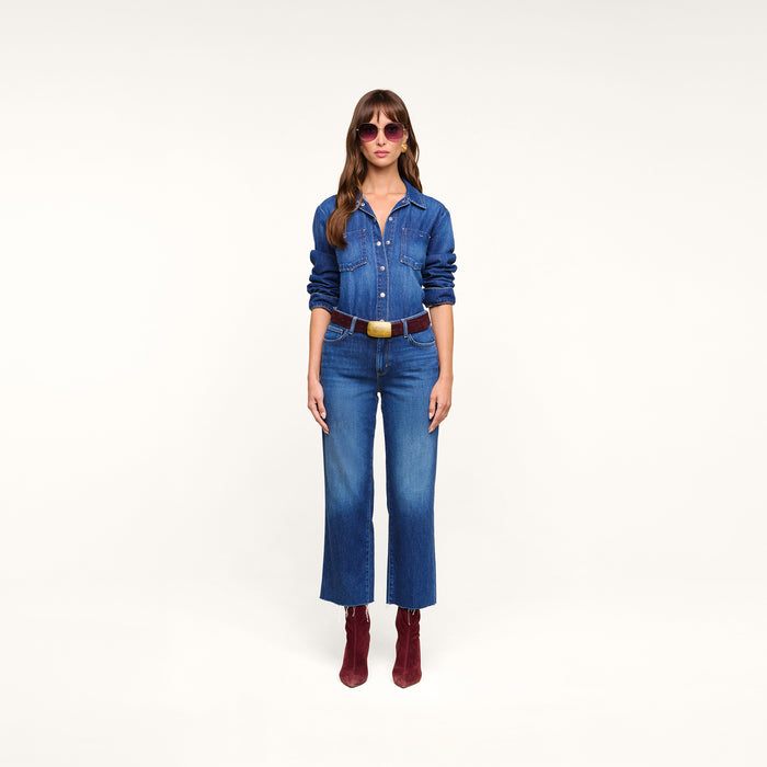 A woman stands facing forward, wearing sunglasses, a blue featherweight denim shirt, the Wanda Cropped Wide-Leg Jean, a brown belt, and red ankle boots, set against a plain white background.