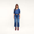 A woman stands facing forward, wearing sunglasses, a blue featherweight denim shirt, the Wanda Cropped Wide-Leg Jean, a brown belt, and red ankle boots, set against a plain white background.