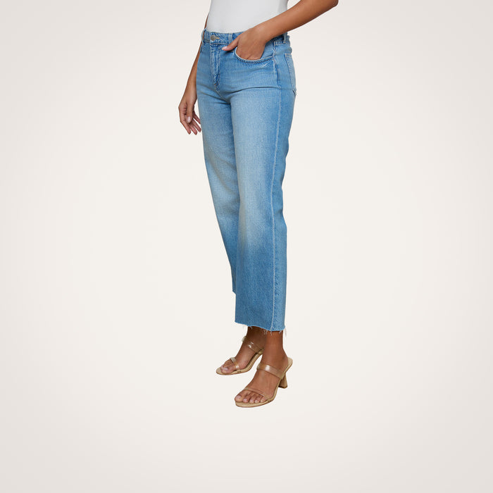 A woman stands against a plain light background, wearing the Wanda Cropped Wide-Leg Jean—light blue, high-waisted, all-cotton denim jeans featuring raw hems and a cropped length—paired with a white top and tan heeled sandals.