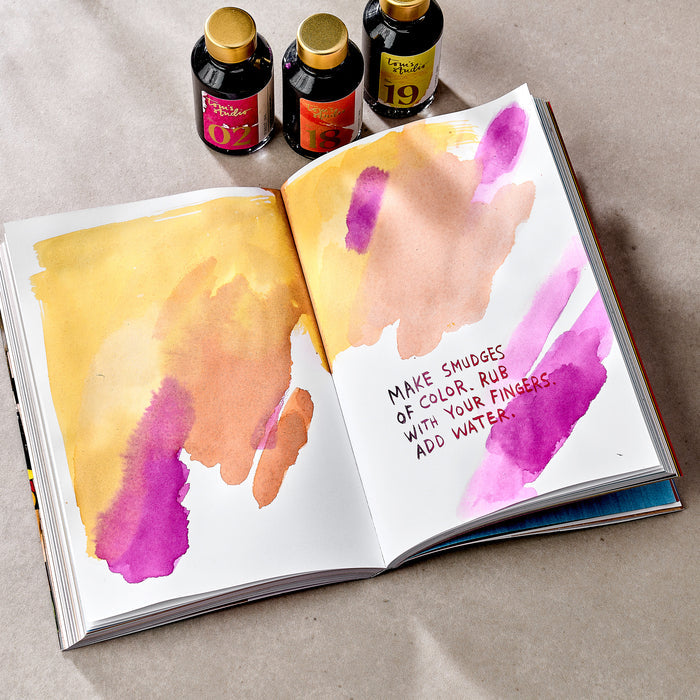 An open spread from Wreck This Journal, featuring colourful smudges of yellow, orange, pink, and purple ink on both pages. The right page includes the handwritten instruction: “MAKE SMUDGES OF COLOR. RUB WITH YOUR FINGERS. ADD WATER.” Three bottles of ink with gold caps are placed above the journal, each labelled with different numbers and colours.