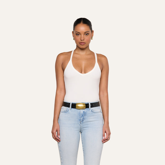 A woman stands facing forward against a plain light background, wearing the Wrenna Racerback Bodysuit in white rib fabric, paired with light blue jeans and a black belt with a large gold buckle.