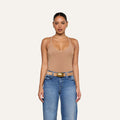A woman stands facing forward against a plain light background, wearing the Wrenna Racerback Bodysuit in tan rib fabric, blue jeans, a tan belt with a gold buckle, and gold hoop earrings.