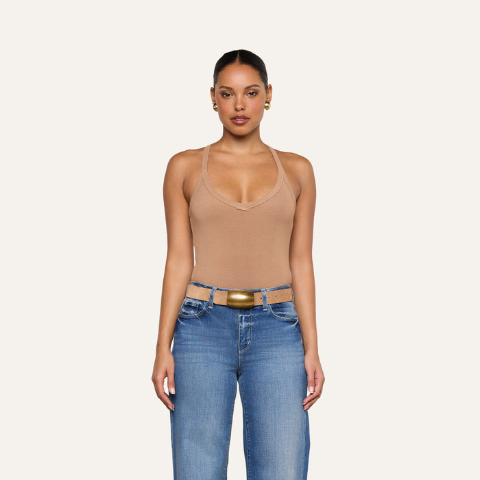 A woman stands facing forward against a plain light background, wearing the Wrenna Racerback Bodysuit in tan rib fabric, blue jeans, a tan belt with a gold buckle, and gold hoop earrings.