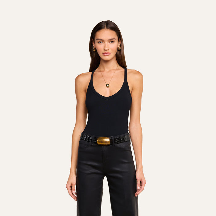 A woman stands against a plain background, wearing the Wrenna Racerback Bodysuit in black rib fabric, paired with black pants featuring a textured belt and large buckle. She accessorizes with gold earrings and a pendant necklace, looking directly at the camera with a neutral expression.