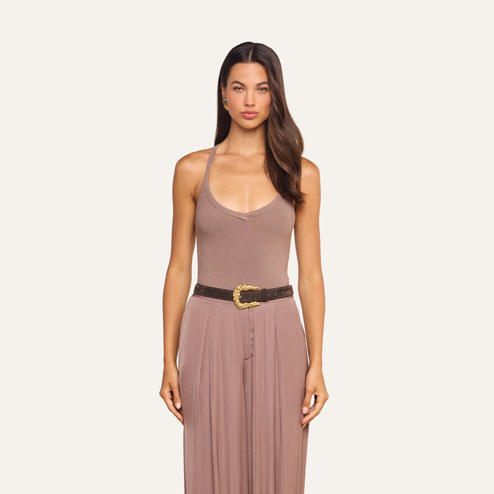 A woman stands facing forward, wearing the Wrenna Racerback Bodysuit in taupe, featuring ribbed fabric and a sleeveless design, styled with a dark brown belt with a gold buckle against a plain light background.