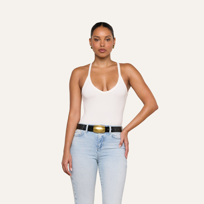 A woman stands against a plain background wearing the Wrenna Racerback Bodysuit in white ribbed fabric, paired with light blue jeans and a black belt with a gold buckle. Her hair is pulled back and one hand rests on her hip.