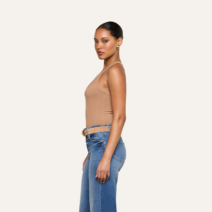 A woman stands in profile against a plain background, wearing the Wrenna Racerback Bodysuit in tan rib fabric, paired with blue jeans and a beige belt; her hair is pulled back and she accessorizes with gold hoop earrings.