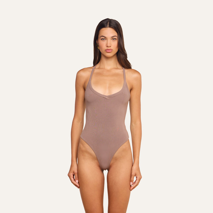 A woman stands facing forward against a plain light background, wearing the Wrenna Racerback Bodysuit in taupe rib fabric.