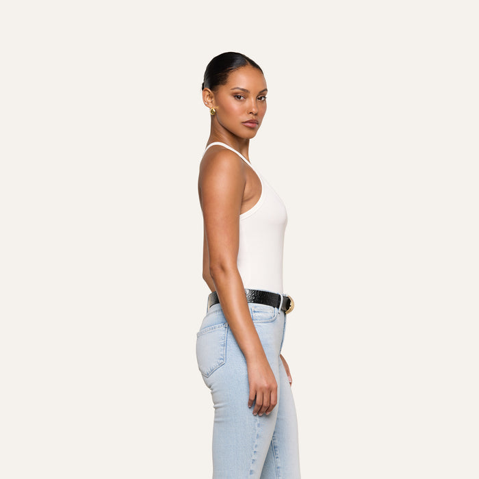A woman stands sideways against a plain light background, her hair pulled back neatly. She is wearing the Wrenna Racerback Bodysuit in white, paired with light blue jeans, a black belt, and gold hoop earrings.