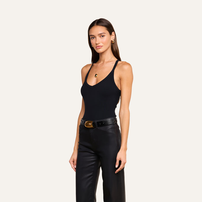 A woman with long brown hair wearing the Wrenna Racerback Bodysuit made of black rib fabric, paired with black leather pants, a black belt with a gold buckle, and gold jewelry, stands against a plain light background.