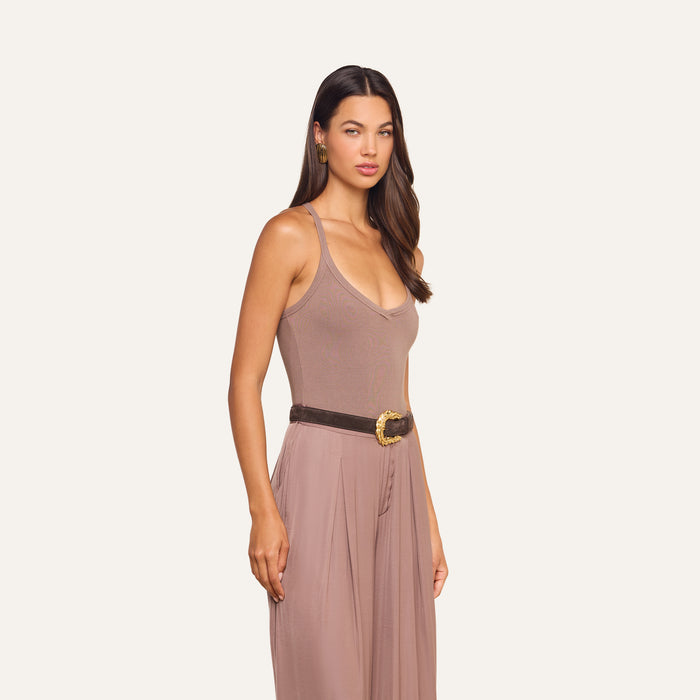 A woman with long brown hair wears the Wrenna Racerback Bodysuit, a taupe sleeveless piece with a V-neckline and subtle ribbed fabric, styled with a dark belt featuring a decorative buckle, as she stands against a plain light background.