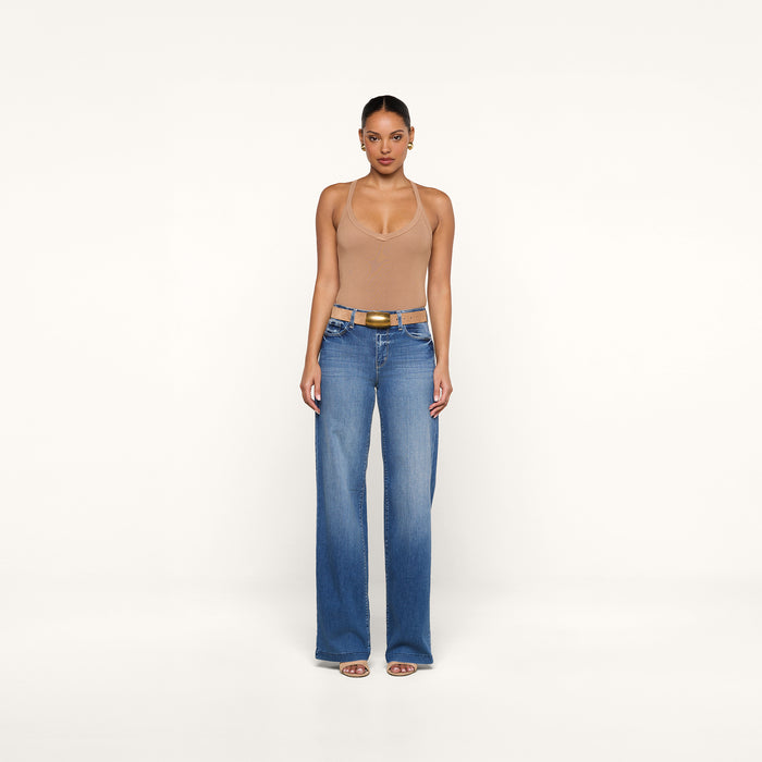 A person stands facing forward, wearing the Wrenna Racerback Bodysuit in tan, paired with blue wide-leg jeans, a brown belt, and sandals against a plain white background.