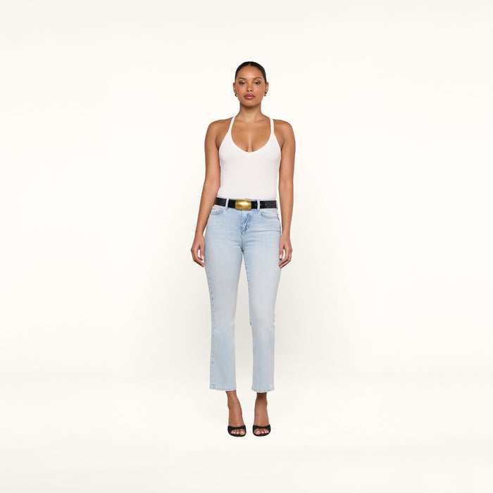 A woman stands against a plain white background, wearing the Wrenna Racerback Bodysuit in white rib fabric, paired with light blue jeans, a black belt with a large buckle, and black open-toe heels. She faces forward with a neutral expression.