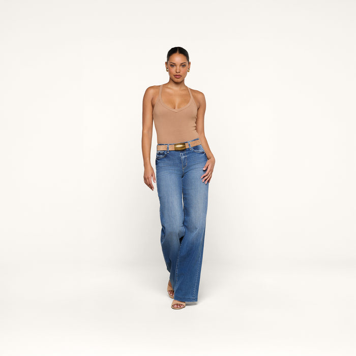 A person stands against a plain white background, wearing the Wrenna Racerback Bodysuit in beige, blue high-waisted wide-leg jeans, a belt, and sandals.