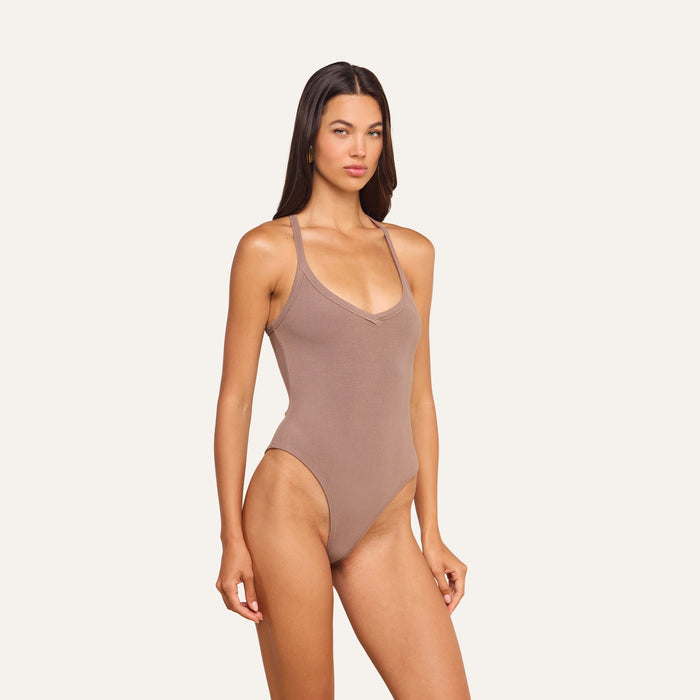 A woman stands confidently against a plain, light background wearing the Wrenna Racerback Bodysuit, featuring taupe-colored ribbed fabric, a V-neckline, and high-cut legs.