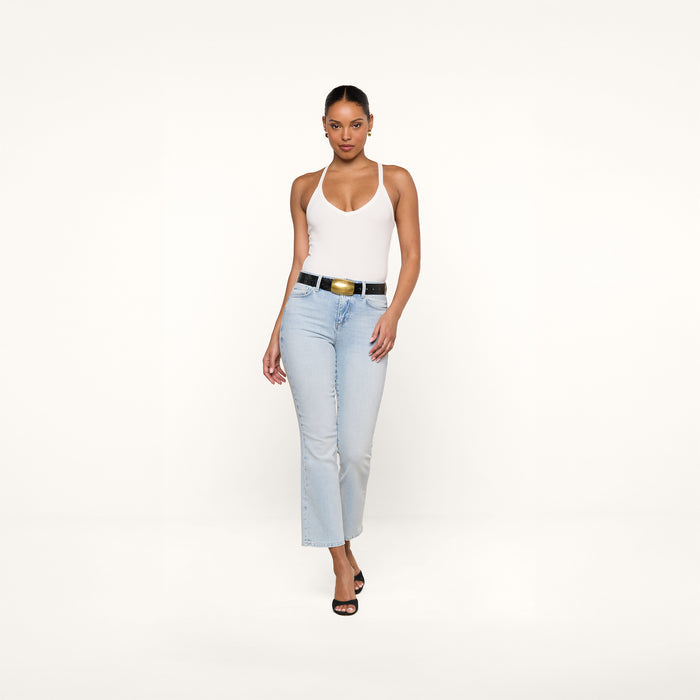 A woman stands against a plain white background, wearing the Wrenna Racerback Bodysuit in white, paired with light blue jeans, a black belt featuring a gold buckle, and black heeled sandals.