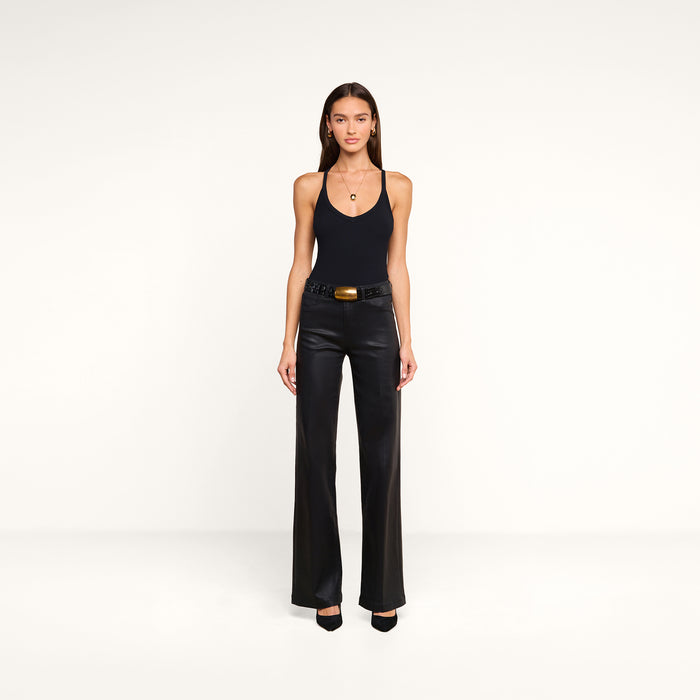 A woman stands against a plain white background, wearing the Wrenna Racerback Bodysuit in black rib fabric, paired with black leather pants, a black belt with a large buckle, and black heels. Her long straight hair frames her neutral expression.