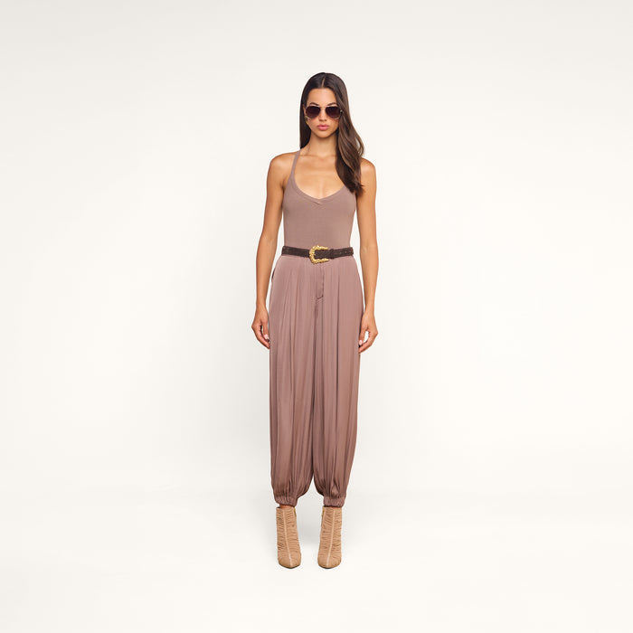 A woman wearing sunglasses, the Wrenna Racerback Bodysuit in taupe rib fabric, loose mauve pants with a belt, and beige boots stands against a plain white background.
