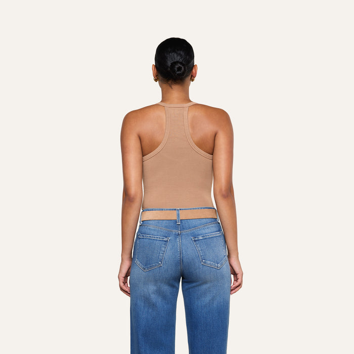 A person with their hair in a bun, wearing the Wrenna Racerback Bodysuit in tan rib fabric and blue jeans, stands facing away from the camera against a plain, light background.