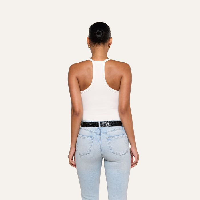 A person with hair in a bun, wearing the Wrenna Racerback Bodysuit in white rib fabric, paired with light blue jeans and a black belt, stands with their back to the camera against a plain, light background.