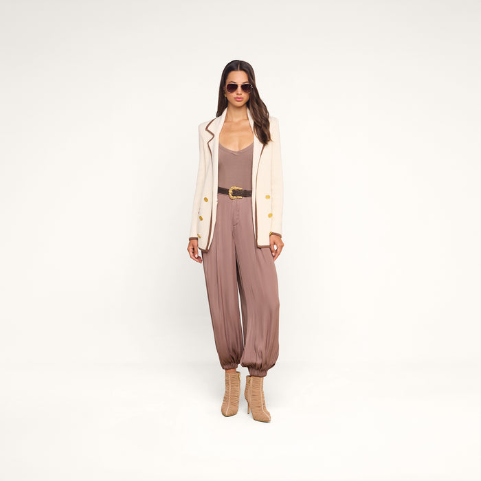 Woman wearing sunglasses, a cream blazer layered over the Wrenna Racerback Bodysuit in taupe rib fabric, brown belt, loose mauve pants gathered at the ankles, and tan heeled boots stands against a plain white background.