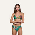 A woman with long brown hair stands with her arms crossed against a plain light background, wearing the Yazmin Bikini Top—a green bikini top featuring a butterfly floral print—and blue earrings.