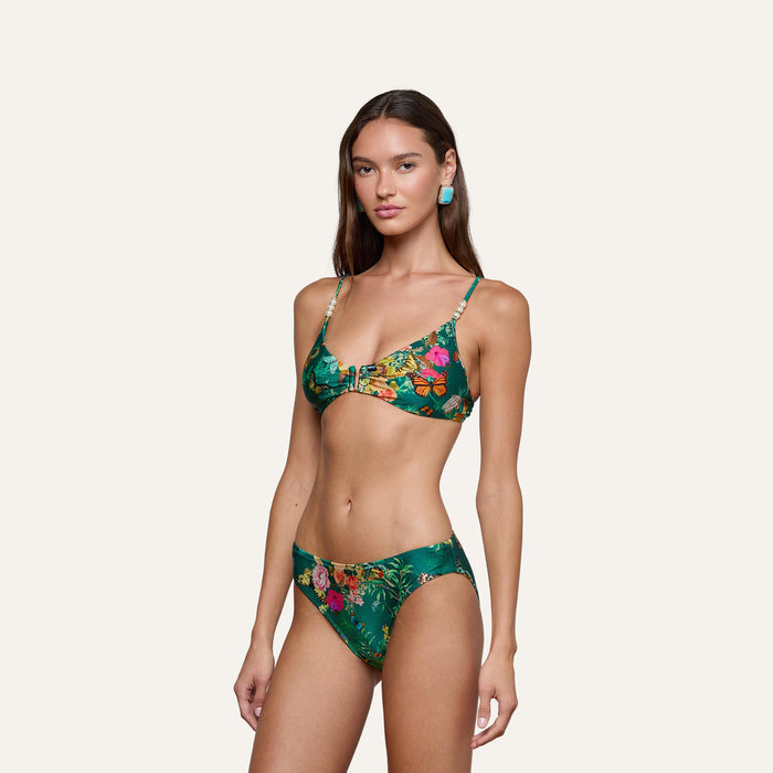 A woman poses in the Yazmin Bikini Top—a green butterfly floral print style featuring a knotted design—paired with matching high-waisted bottoms. She has long brown hair and turquoise earrings, standing against an off-white background.