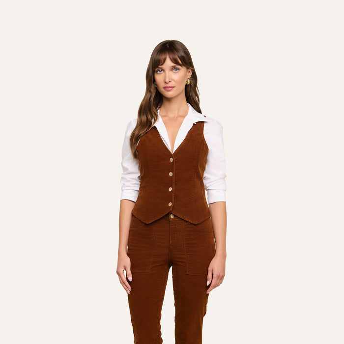 A woman stands against a plain light background, facing forward with a neutral expression, wearing a white collared shirt under the tailored Yelena Vest and matching brown stretch corduroy pants.