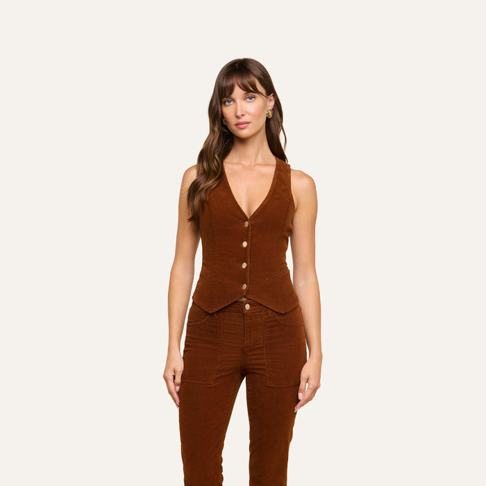 A woman with long brown hair and bangs stands against a plain background, wearing the Yelena Vest in brown stretch corduroy paired with matching brown pants. Her expression is neutral.