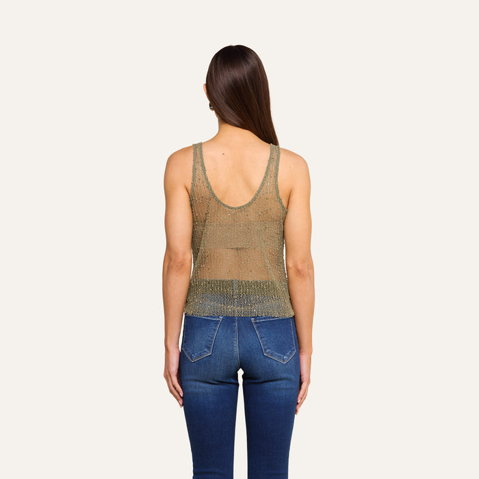 A person with long brown hair, wearing the sleeveless, glittery gold Zia Tank and blue jeans, is standing facing away from the camera against a plain light background.